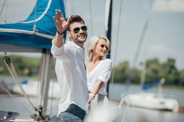Happy young man in sunglasses smiling at camera and waving hand while holding hands with beautiful...