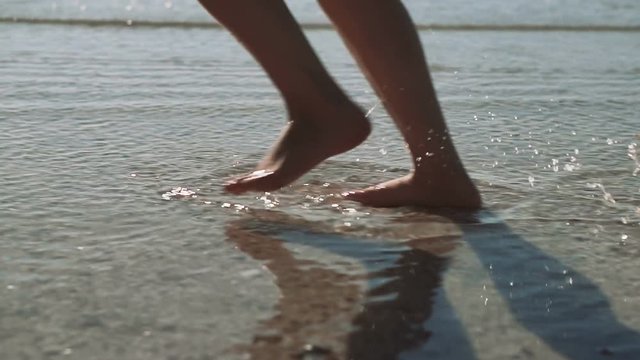 Legs of a young woman walking along the seacoast. Woman runs along the coast splashing sea water, slow motion. Woman runs barefoot into shallow transparent sea water.