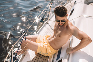 shirtless muscular man in swim trunks and sunglasses having sunbath on yacht