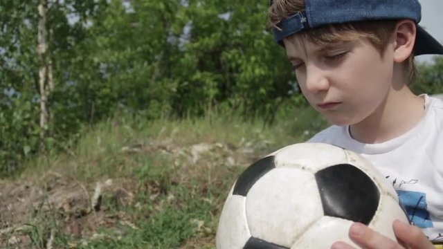 7 year-old Russian white boy in cap with closed eyes and a soccer ball in his hands praying for football team on the background of nature green trees. Football children education concept. Slow motion