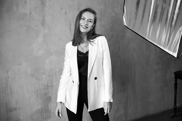 Young beautiful pretty woman with long straight hair wearing black blouse and trousers and white striped jacket laughing, smiling and posing and standing against gray wall