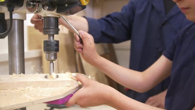 Teenager boy drills a hole on a wooden plank using industrial equipment in carpentry workshop