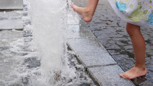 Barefoot child girl legs play with the city street fountain water jets in hot day