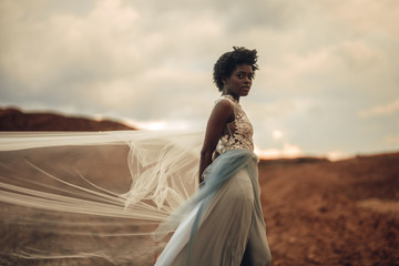 Fototapeta na wymiar Black bride in waving long wedding dress and bridal veil stands on background of beautiful landscape.