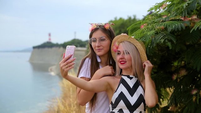 Two positive girls blonde in hat and brunette with glasses fooling around and make selfie pictures with phone standing near green bush tree at rocky cliff by sea on blurred background of lighthouse