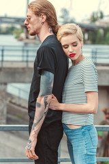 stylish girlfriend hugging tattooed boyfriend from back on bridge