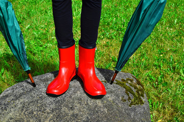Teen girl legs wearing red rubber boots standing on big gray stone with two umbrellas on green...