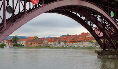Zabytkowe, malownicze centrum Mariboru, Słowenia, widok spod żelaznego, różowego mostu (przęsła), rzeka Sawa, budynki z czerwonymi dachami, zaparkowane samochody, nabrzeże
