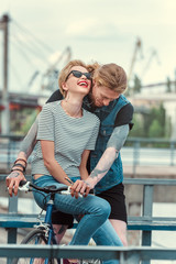 tattooed boyfriend and smiling stylish girlfriend on bicycle on bridge