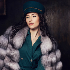 Stylish woman wearing green jacket dress and cap, fur coat and black jackboots posing in dark...