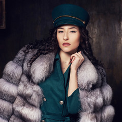 Stylish woman wearing green jacket dress and cap, fur coat and black jackboots posing in dark...