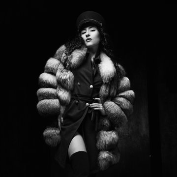 Stylish woman wearing green jacket dress and cap, fur coat and black jackboots posing in dark interior. Fashion style portrait. Brunette woman with curly hair. Full length portrait.