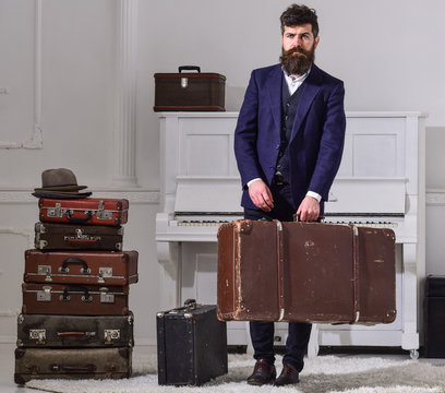 Macho elegant on strict face stands near pile of vintage suitcase, holds suitcase. Man, traveller with beard and mustache with luggage, luxury white interior background. Baggage delivery concept.