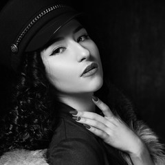 Stylish woman wearing green jacket dress and cap, fur coat and black jackboots posing in dark interior. Fashion style portrait. Brunette woman with curly hair. Close-up portrait.