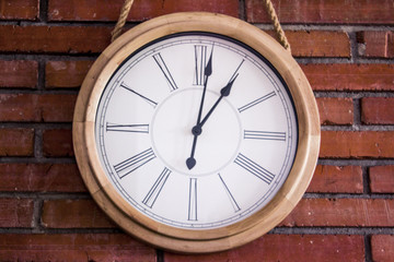Fototapeta na wymiar Close-up of a wooden classic and simple wall clock hanging in a red brick wall by a rope. It has roman numerals. Eye-level shot.