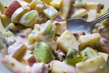 Detail close-up of a plate with chopped apples and kiwis mixed with granola and yogurt. A steel spoon rest in the fruit.