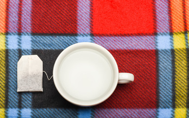 Mug filled with boiling water and teabag on colorful plaid background. Tea time concept. Process of tea brewing in ceramic mug, top view. Cup or white mug with transparent hot water and bag of tea