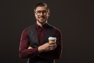 stylish buisnessman in eyeglasses holding disposable coffee cup and smiling at camera isolated on black