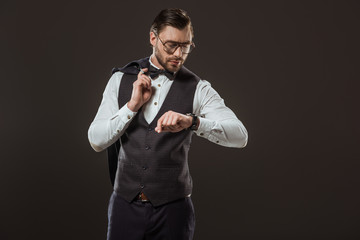 stylish businessman in bow tie and eyeglasses holding suit jacket and checking wristwatch isolated on black
