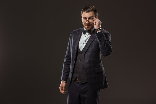 portrait of handsome smiling man in suit and bow tie adjusting eyeglasses isolated on black