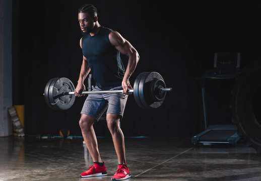 athletic african american man in sportswear lifting barbell and looking away in gym - Powered by Adobe