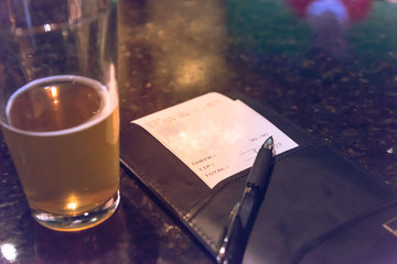 Vintage open pint glass beer next to leather bill holder with restaurant check and pen. Close-up,...