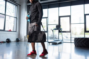 cropped shot of african american man in sportswear holding bag and bottle of water in gym
