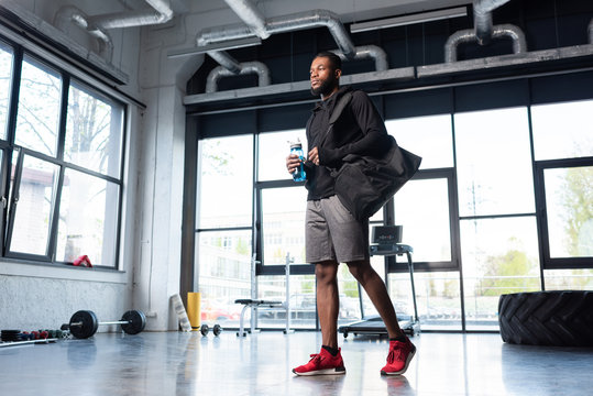 low angle view of sportive african american man holding bottle of water and looking away in gym
