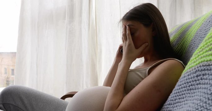 Side view of young woman in expectancy sitting on sofa at home and having phone call looking sad