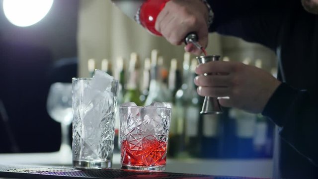 person with bottle into arms measures proportion of liquor in jigger and pours it into glass with ice in bar close up