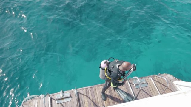 Unrecognized bald scuba diver fools around, uses a flipper as a musician guitar, before diving under the water on an ocean yacht. Slow motion.