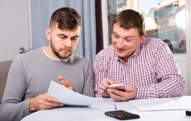 Two cheerful men with documents and phone at home table