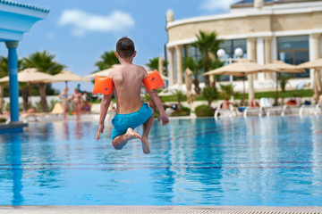 Naklejka premium European boy in floating sleeves having fun jumping into swimming pool at resort. Back view.