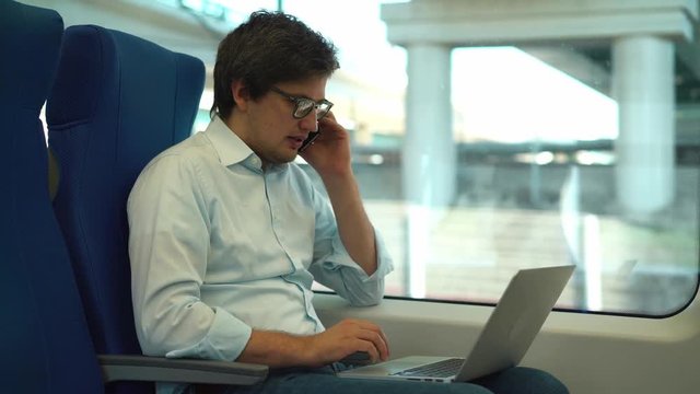 Handsome businessman in a shirt and jeans is talking on the phone and using his laptop while riding a train. Handheld real time medium shot