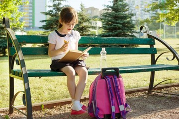Portrait of schoolgirl 7 years old on a bench reading book, eating ice cream. Background school yard
