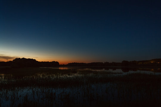 A bright sunset in the night sky. The landscape with the river and trees is photographed on a long exposure.