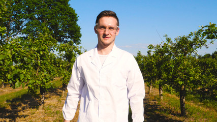 A young handsome (male) biologist or agronomist, wearing a white coat, wearing goggles, wearing blue rubber gloves, walks across the apple tree, the background of nature and greenery.