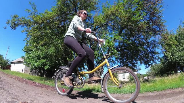 Young Woman Riding Vintage Bicycle along a Rural Road in a Village. The Camera moves low over the ground. Active woman spending her weekend outdoors riding old bike. Steady shot. Summer day, Blue sky.