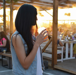 Portrait of a young beautiful dark-haired woman in a blue denim vest drinking orange juice in a summer amusement park. Attractive. Summer. Fresh. Lifestyle. Sunset. Sunlight.