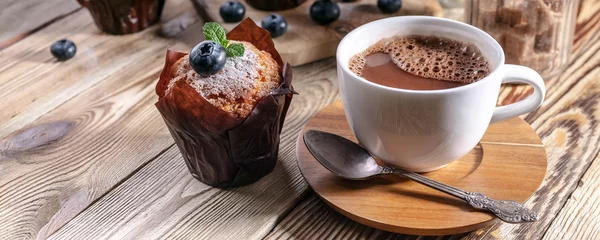  Muffins met bosbessen en een kop warme chocolademelk op een houten ondergrond. zelfgemaakt bakken. Banner © yusev
