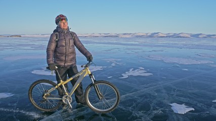 Fototapeta premium Man and his bicycle on ice. He stand and looks at the beautiful sunset. The cyclist is dressed in a gray down jacket, backpack and helmet. Ice of the frozen Lake Baikal. The tires on the bicycle are