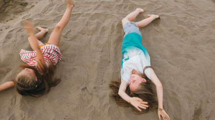 Two teenage are lie on a sandy beach near the sea. Girl playing, talk to each other. Sisters are dressed in dresses and sunglasses. Children have real emotions of happiness.