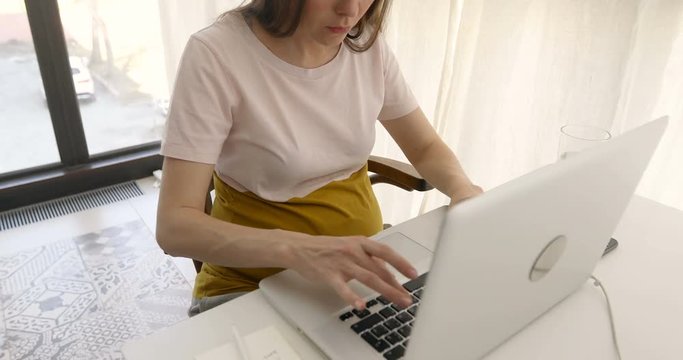 Crop casual pregnant woman sitting at workplace using laptop and taking notes