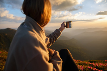woman traveler drinks coffee with a view of the mountain landscape. A young tourist woman drinks a hot drink from a cup and enjoys the scenery in the mountains. Trekking concept - 210272908