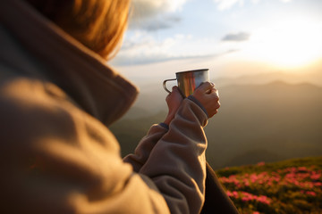 Closeup photo of cup with tea in traveler's hand over out of focus mountains view. A young tourist woman drinks a hot drink from a cup and enjoys the scenery in the mountains. Trekking concept - 210272915