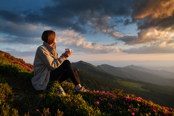 woman traveler drinks coffee with a view of the mountain landscape. A young tourist woman drinks a hot drink from a cup and enjoys the scenery in the mountains. Trekking concept - 210272949