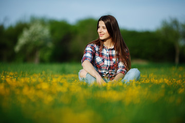 Pretty woman in plaid shirt and jeans sitting on the grass in the meadow of flowers. Pretty girl relaxing outdoors on spring grass