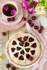 A cherry pie decorated with heart-shaped ornamentation with a cup of tea, a bouquet of flowers and ripe cherry berries.