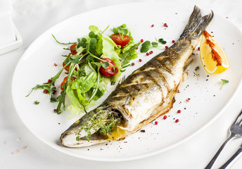 Baked fish with lemon and fresh vegetables on a white plate in the restaurant