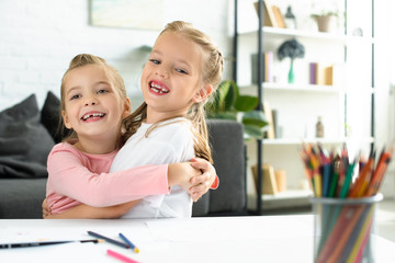 portrait of happy little children hugging each other at table with papers and pencils for drawing at home